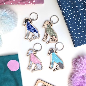 May include: Four wooden dog keyrings with silver rings. Each dog is a different colour, including blue, green, pink, and blue, and has a patterned sweater. The keyrings are on a white surface with fabric and pom-poms.