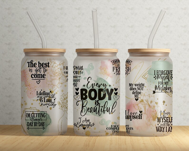 May include: Three frosted glass tumblers with bamboo lids and clear straws. Each tumbler features inspirational quotes and floral designs in gold, pink, and green. The text includes phrases like "Every Body is Beautiful" and "I love myself."