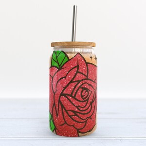May include: A stainless steel tumbler with a wooden lid and a straw. The tumbler has a red glitter rose design with green leaves.