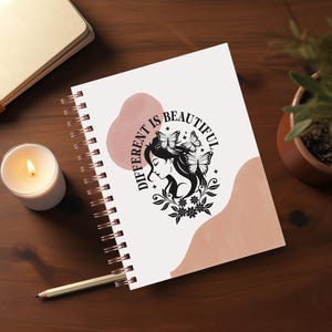 May include: A white spiral bound notebook with a pink and white abstract design. The notebook has the text "Different is Beautiful" with a black silhouette of a woman with butterflies in her hair.