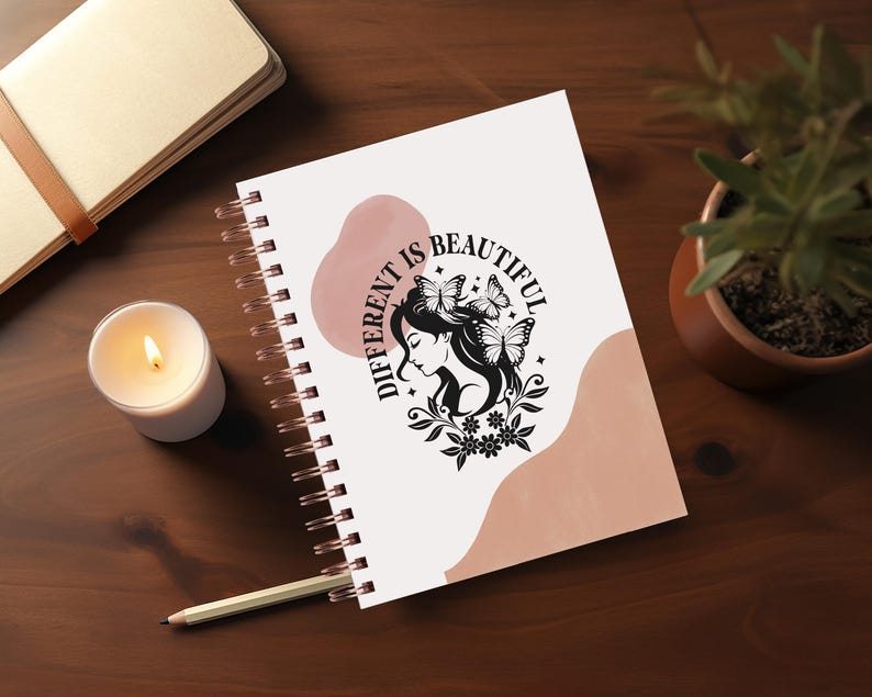 May include: A white spiral bound notebook with a pink and white abstract design. The notebook has the text "Different is Beautiful" with a black silhouette of a woman with butterflies in her hair.