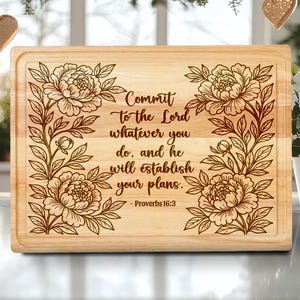 May include: A rectangular wooden cutting board with a floral design and the text "Commit to the Lord whatever you do, and he will establish your plans. Proverbs 16:3" engraved on it. The board has a light brown color.
