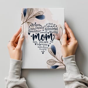 May include: A white notebook with the word "Mom" in the center, surrounded by words like "Brave," "Smart," and "Beautiful." Floral and leaf accents in blue and beige decorate the cover. The notebook is held by two hands.