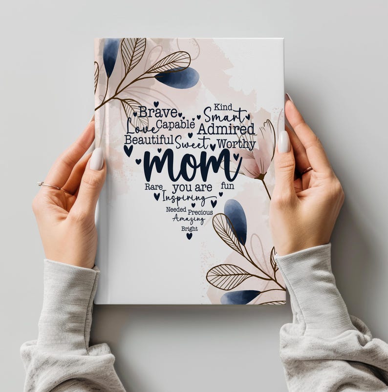 May include: A white notebook with the word "Mom" in the center, surrounded by words like "Brave," "Smart," and "Beautiful." Floral and leaf accents in blue and beige decorate the cover. The notebook is held by two hands.