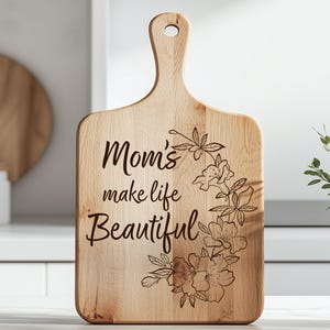 K&ouml;nnte beinhalten: Holzschneidebrett mit Griff und dem eingravierten Text "Mom's make life Beautiful". Das Brett hat ein Blumendesign. Das Schneidebrett ist aus hellem Holz gefertigt.