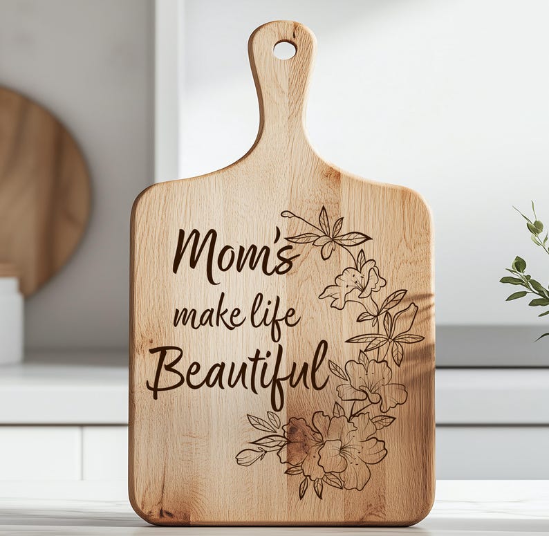 K&ouml;nnte beinhalten: Holzschneidebrett mit Griff und dem eingravierten Text "Mom's make life Beautiful". Das Brett hat ein Blumendesign. Das Schneidebrett ist aus hellem Holz gefertigt.