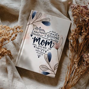May include: A hardcover journal with the word "Mom" in the center, surrounded by words like "Brave," "Love," and "Inspiring." The cover features floral and leaf designs in blue and pink, with a cream-colored background.