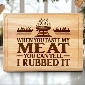 May include: A rectangular wooden cutting board with a carved design. The design includes a grill, flames, and the text "WHEN YOU TASTE MY MEAT - YOU CAN TELL - I RUBBED IT". The board has a light brown color and a groove around the edge.