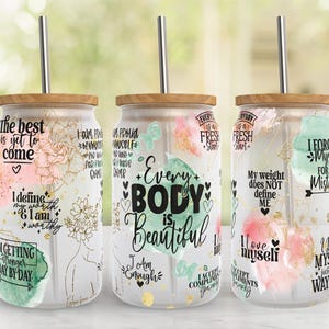 May include: Three glass tumblers with wooden lids and metal straws. Each tumbler features inspirational quotes and floral designs in pink, gold, and green. The text includes phrases like "Every Body is Beautiful" and "The best is yet to come."