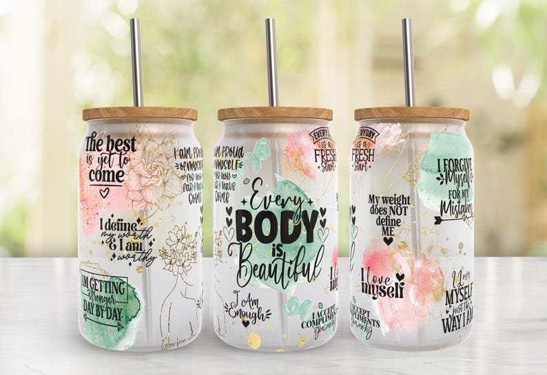 May include: Three glass tumblers with wooden lids and metal straws. Each tumbler features inspirational quotes and floral designs in pink, gold, and green. The text includes phrases like "Every Body is Beautiful" and "The best is yet to come."
