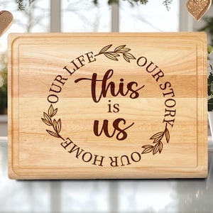 May include: A rectangular wooden cutting board with a juice groove. The board features the text "This is us" inside a circular wreath, with the words "Our Life, Our Story, Our Home" around the circle. The wood has a light brown color.