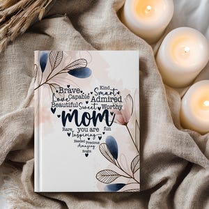 May include: A white journal with the word "Mom" in a heart-shaped font, surrounded by words like "Brave," "Love," and "Inspiring." Floral and leaf designs in blue and pink accent the cover. The journal is on a beige cloth.