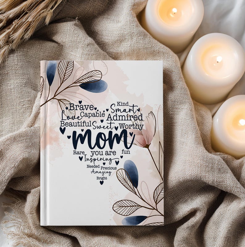 May include: A white journal with the word "Mom" in a heart-shaped font, surrounded by words like "Brave," "Love," and "Inspiring." Floral and leaf designs in blue and pink accent the cover. The journal is on a beige cloth.