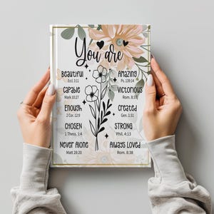 Op de afbeelding: Een rechthoekig notitieboek met een witte kaft, een gouden rand, zachte perzikkleurige bloemillustraties en zwarte tekst: You are Beautiful, Amazing, Capable, Victorious, Enough, Created, Chosen, Strong, Never Alone, Always Loved.