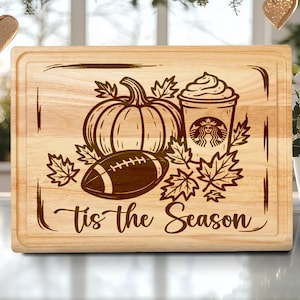 May include: A light-colored wooden cutting board with a carved design featuring a pumpkin, football, coffee cup, and fall leaves. The text "Tis the Season" is also carved into the board.