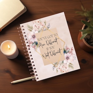 May include: A pink and white floral design journal with a gold spiral binding. The journal has a quote on the front that reads "You've got to move different if you want different."