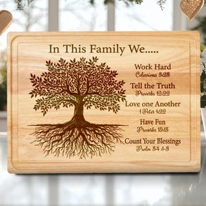 May include: A rectangular wooden cutting board with a carved tree design and the text "In This Family We..." followed by inspirational phrases and Bible verses. The board has a light wood grain.