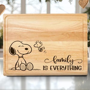 May include: A rectangular wooden cutting board with a light brown finish. The board features a black illustration of Snoopy and Woodstock, with the text "family IS EVERYTHING" in a cursive font. The board has a recessed edge.