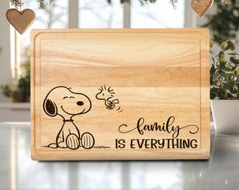 Può includere: Tagliere rettangolare in legno chiaro. Presenta un'illustrazione nera di Snoopy e Woodstock, con la scritta "family IS EVERYTHING" in corsivo. Il tagliere ha un bordo rientrante.
