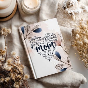 May include: A white hardcover journal with the word "Mom" in a script font, surrounded by floral and leaf illustrations. The journal includes words like "Brave," "Smart," and "Inspiring." The journal is on a neutral-toned fabric.