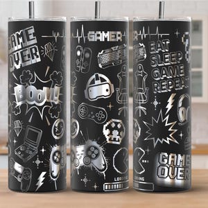 May include: Black stainless steel tumbler with a silver gamer-themed design. The design includes game controllers, headsets, and the words "Game Over," "Boom!," and "Eat Sleep Game Repeat." The tumbler has a silver straw.