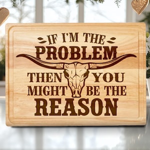 May include: A rectangular wooden cutting board with a carved design. The design features a longhorn skull and the text "IF I'M THE PROBLEM THEN YOU MIGHT BE THE REASON" in a dark brown color. The board has a juice groove around the edge.