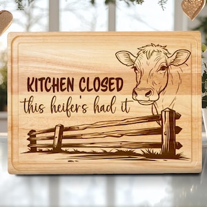 May include: A rectangular wooden cutting board with a carved design. The design features a cow's head, a wooden fence, and the text "KITCHEN CLOSED this heifer's had it". The board has a light brown color and a beveled edge.