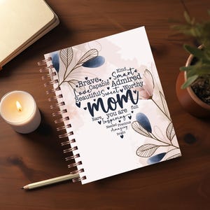 May include: A spiral-bound notebook with the word "Mom" in a decorative font, surrounded by words like "Brave" and "Sweet." The notebook has floral accents in blue and pink. A lit candle and a pencil are next to the notebook.