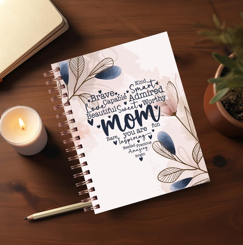May include: A spiral-bound notebook with the word "Mom" in a decorative font, surrounded by words like "Brave" and "Sweet." The notebook has floral accents in blue and pink. A lit candle and a pencil are next to the notebook.