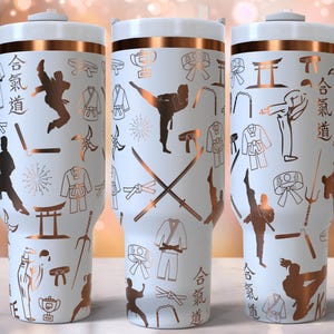 Puede incluir: Taza de viaje blanca con tapa de color cobre y un diseño blanco y cobre. El diseño presenta una variedad de símbolos y figuras de artes marciales, incluyendo una postura de karate, una espada y un nunchaku. El texto "合気道" está impreso en la taza.