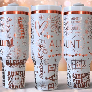 May include: Three white insulated tumblers with rose gold detailing and handles. Each tumbler is decorated with the word "Aunt" and related phrases, along with floral and heart designs. The tumblers have a white lid and a rose gold band.