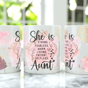 以下が含まれることがあります： ピンクの水彩アクセントと金色のフローラルデザインが施された白いセラミックマグ。マグには「She is」の文字と「強い、恐れを知らない、温かい、愛情深い、忍耐強い、無私の、おば」などの説明的な言葉が書かれています。