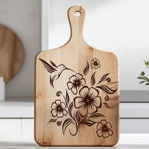 K&ouml;nnte beinhalten: Ein h&ouml;lzernes Schneidebrett mit Griff und einem dekorativen Kolibri- und Blumendesign. Der Kolibri ist im Flug dargestellt, mit Blumen und Bl&auml;ttern, die in Dunkelbraun ge&auml;tzt sind. Das Brett ist aus hellem Holz gefertigt.