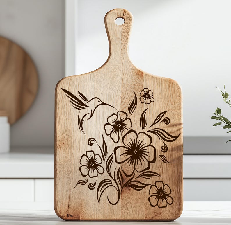 K&ouml;nnte beinhalten: Ein h&ouml;lzernes Schneidebrett mit Griff und einem dekorativen Kolibri- und Blumendesign. Der Kolibri ist im Flug dargestellt, mit Blumen und Bl&auml;ttern, die in Dunkelbraun ge&auml;tzt sind. Das Brett ist aus hellem Holz gefertigt.