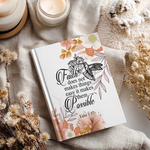 Puede incluir: Un diario blanco con la cita "La fe no hace las cosas fáciles, las hace posibles" e ilustraciones florales en rosa, melocotón y blanco. Un colibrí está representado en el diseño. El diario está rodeado de flores secas y velas.
