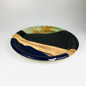 May include: Oval ceramic serving platter with a dark blue rim and a painted design. The design includes a black section, a tan section, and a green section with orange accents. The platter is on a white background.