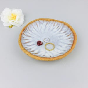 Peut inclure: Un vide-poche en céramique ronde avec un bord beige texturé non émaillé et un centre émaillé blanc présentant un motif radial en forme de pétales. À l'intérieur se trouvent une petite perle rouge en forme de cœur et une bague dorée fine.