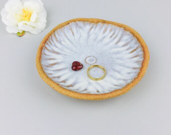 May include: A round ceramic trinket dish with a textured, unglazed tan rim and a white glazed center featuring a radial, petal-like pattern. Resting inside the dish are a small, dark red heart-shaped bead and a simple, thin gold-toned metal ring.