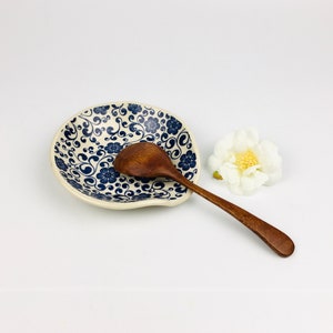 May include: A wooden spoon lies on a blue and white floral patterned spoon rest. The spoon rest is oval-shaped with a raised edge. A white flower with a yellow centre is to the right of the spoon rest.