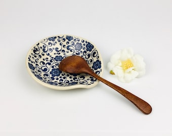 May include: A wooden spoon rests on a blue and white floral patterned spoon rest. The spoon rest is oval-shaped with a raised edge. A white flower with a yellow center is to the right of the spoon rest.