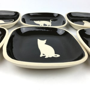 May include: Set of six square ceramic dishes with a glossy black interior and off-white rim. Each dish features a white cat silhouette in various poses. The dishes are ideal for serving appetizers or small portions.