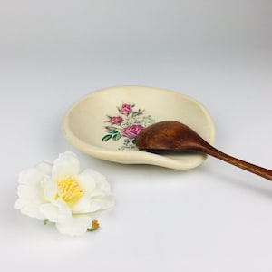 May include: An off-white ceramic spoon rest with a floral rose design and a wooden spoon. The spoon rest has a curved shape with a raised edge. A white flower with a yellow center is in the foreground.