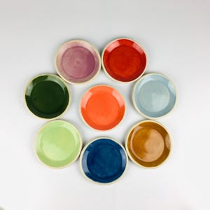 May include: Eight small, round ceramic plates in a variety of colours, including red, orange, green, blue, and purple. Each plate has a white rim and a glossy finish. The plates are arranged in a circular pattern against a white background.