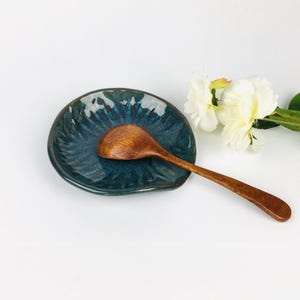 May include: A teal ceramic spoon rest with a textured surface, holding a wooden spoon. The spoon rest has a unique shape and is placed on a white surface. A white flower is in the background.