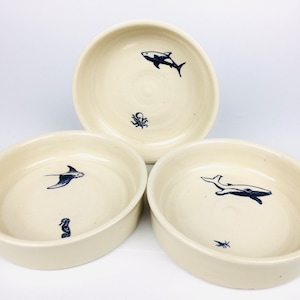 May include: Three off-white ceramic bowls with navy blue sea creature illustrations. One bowl features a killer whale and an octopus, another a manta ray and a seahorse, and the third a whale and a small fish. The bowls are round with a slightly raised edge.