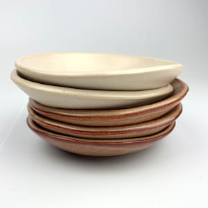 May include: A stack of six handmade ceramic dishes. The oval-shaped dishes have a slightly raised rim. The top dishes are a light beige colour, while the lower dishes have a reddish-brown rim and a tan centre.