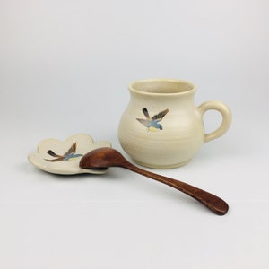 May include: A cream-coloured ceramic mug with a handle and a matching spoon rest, both adorned with a painted bluebird design. A dark wooden spoon rests on the spoon rest. The mug is approximately 10 cm tall.