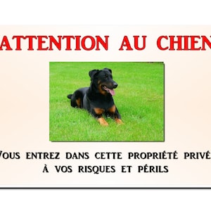 Op de afbeelding: Een rechthoekig bord met de tekst "ATTENTION AU CHIEN" in rode letters. Daaronder een foto van een zwart-bruine hond die op groen gras ligt. Het bord bevat ook de tekst "VOUS ENTREZ DANS CETTE PROPRIÉTÉ PRIVÉE À VOS RISQUES ET PÉRILS".
