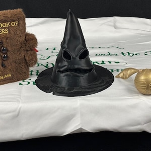 May include: A black Sorting Hat with a face-like design, a golden Snitch, and a brown, furry book titled "The Monster Book of Monsters" are displayed on a white surface. The book has black button eyes and the text "SHEGLAM".