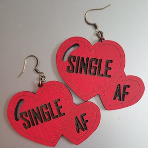 May include: A pair of red wooden earrings shaped like hearts. Each heart has the words "SINGLE AF" written on it in black.